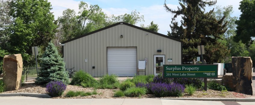Surplus Property Building Front of Building with front landscaping and rocks and parking lot on a sunny day.